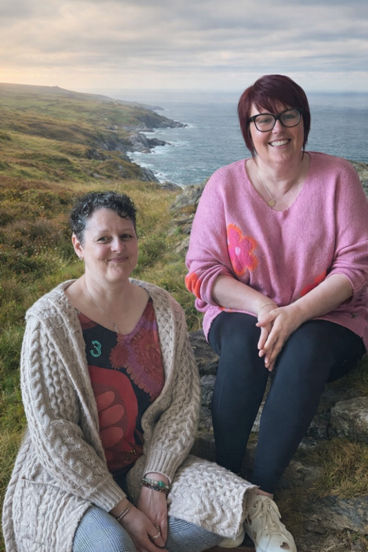 Sharyn and Marie, co-founders of The Exhale Awakening, standing together. North Yorkshire woodland behind Sharyn on the left, Donegal coastline behind Marie on the right.